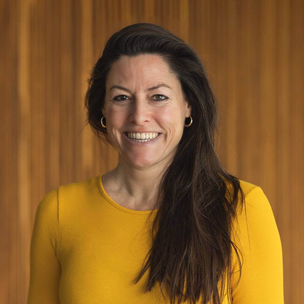 Frau in gelbem Oberteil lächelt vor Holzwand.